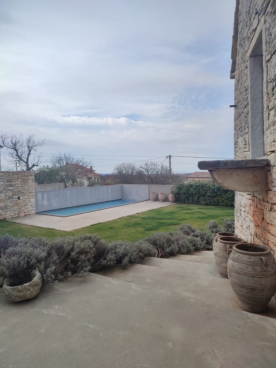 Obalni vrt izvan sezone: bazen za plivanje, obrezana lavanda i terakota posude uz kamenu kuću, stabla iza još gola
